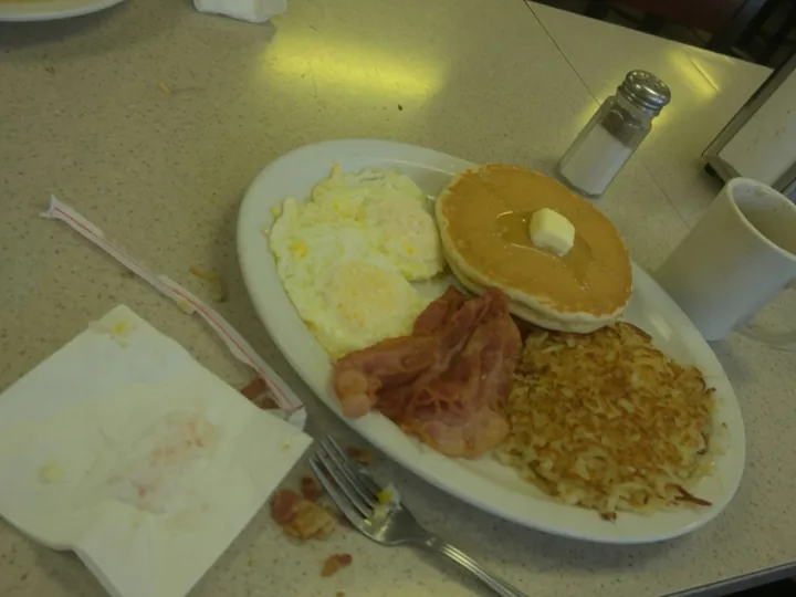 AI-made sample, before: a quick phone photo of a diner breakfast plate, tilted, in dim yellow light, with a used napkin, a straw wrapper and a fork on the counter.